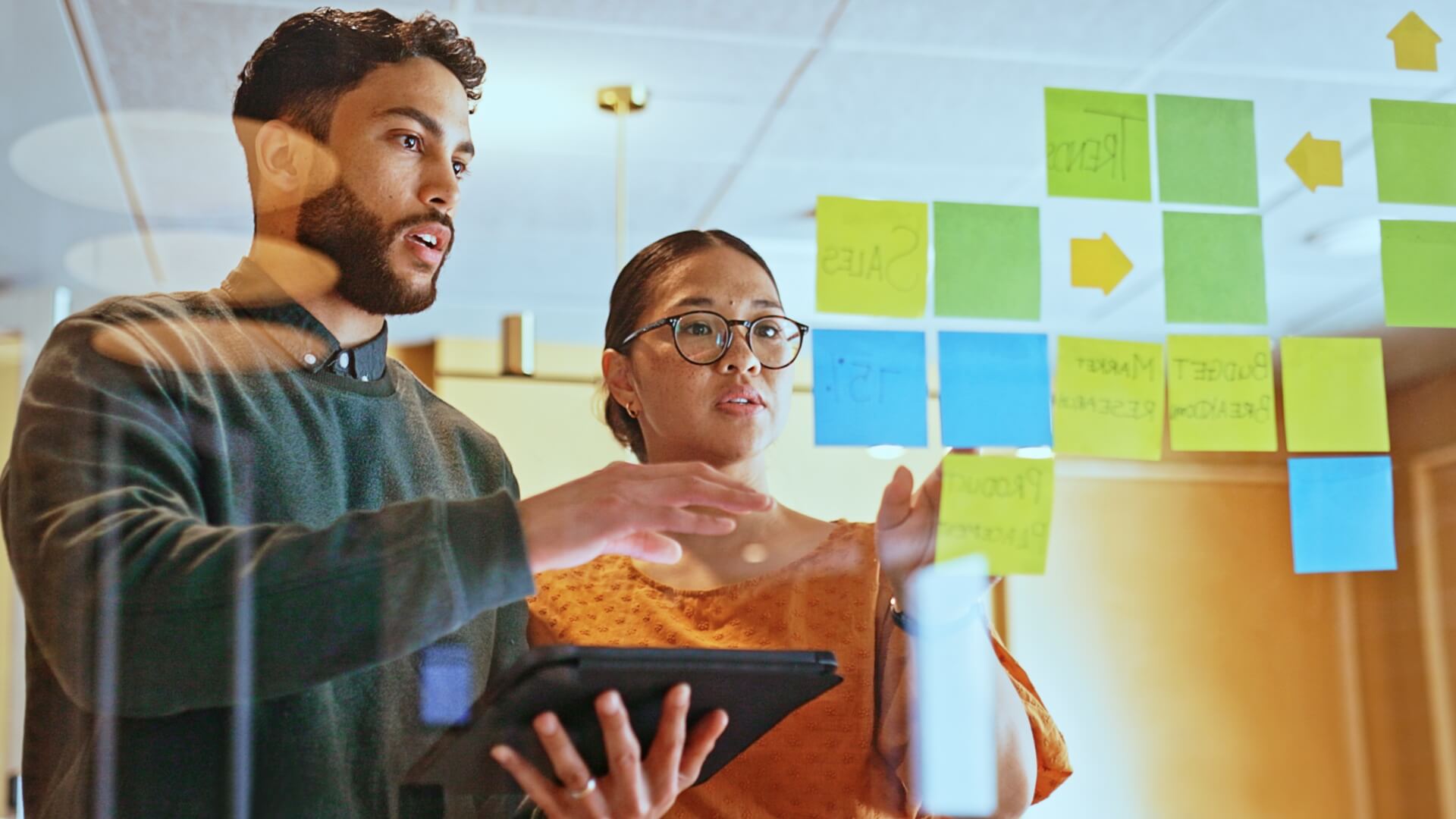 a man and woman planning work strategies with sticky notes on a glass wall in a meeting room