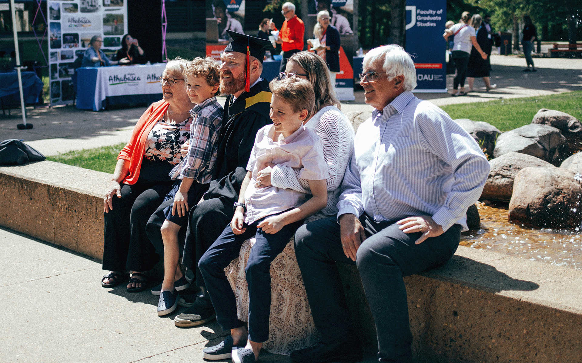 A family surrounds a graduate while sitting near the fountains on Athabasca University campus