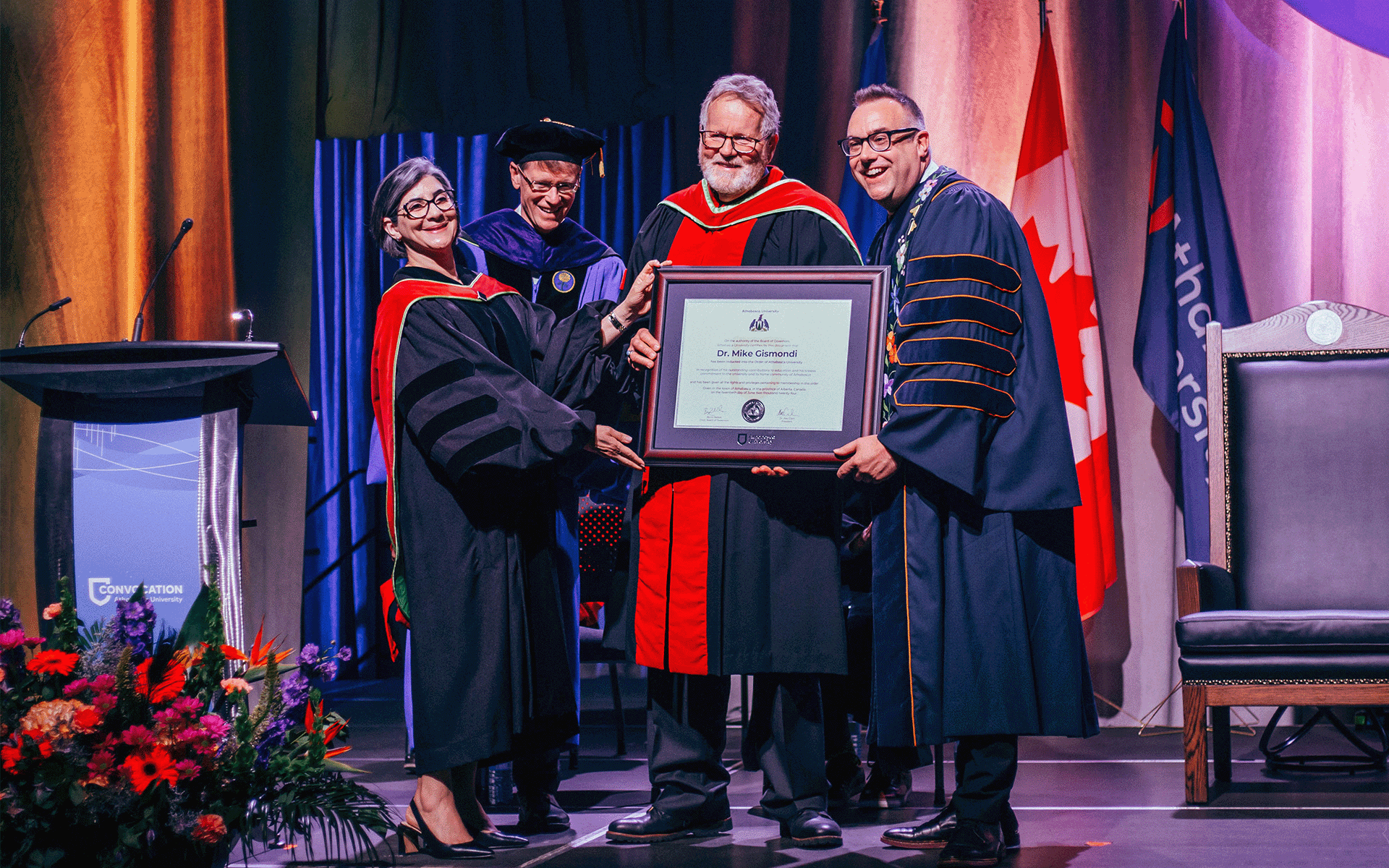 A woman and three men standing together with one of them holding a large framed document for the Order of Athabasca University awarded to Dr. Mike Gismondi during the convocation ceremony