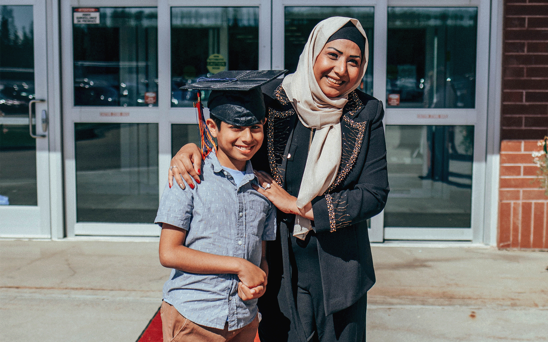 Young boy wearing grad cap and woman standing next to him. Both of them are smiling at the camera