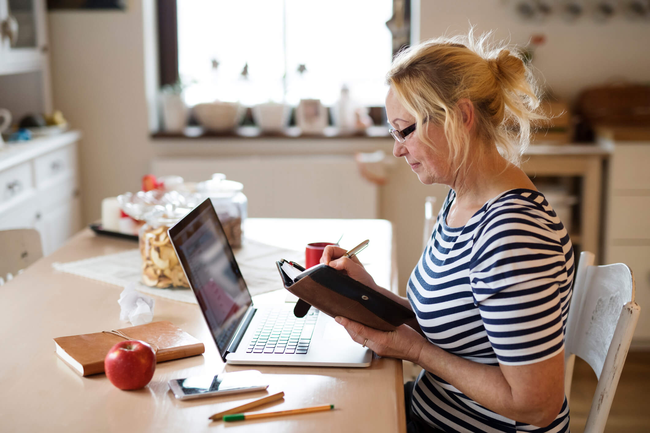 Senior woman with laptop at home at the kitchen table, writing something into her calendar