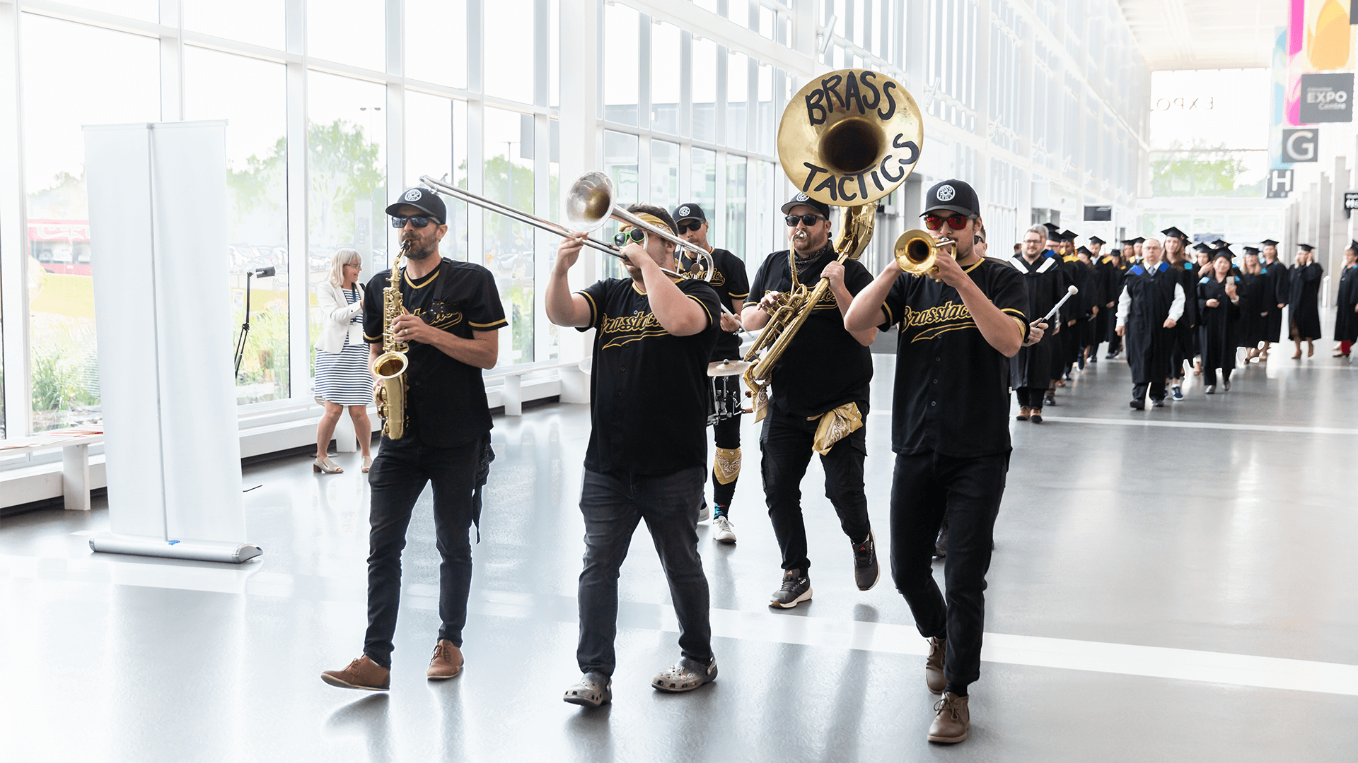 shot of band brass tactics leading the convocation procession. there is a man with a saxophone, a many with a trombone, a man with a trumpet, a man with a tuba